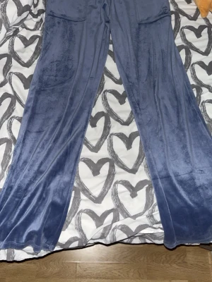 Blå velourbyxor Juicy Couture - Mysiga blå velourbyxor från Juicy Couture med raka ben och fickor framtill. Byxorna har en mjuk och glansig finish som ger en lyxig känsla, perfekta för chill hemma eller som loungewear. Klassisk logga på fickan för en ikonisk touch.