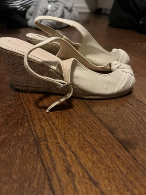 Beigea kilklackar från Nadin - Säljer ett par snygga beige sandaler från Nadin med kilklack i flätad design. Sandalerna har öppen tå, knytdetalj framtill och justerbar rem runt hälen. Perfekta för sommarens alla tillfällen och ger en chic touch till din outfit.