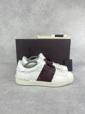 Valentino vita sneakers med nitar - Snygga vita sneakers från Valentino med bred vinröd rem över vristen dekorerad med matchande nitar. Skorna har en klassisk låg siluett, vita snören och detaljerad sula med små nitar baktill. Tillverkade i läder för en lyxig känsla.
