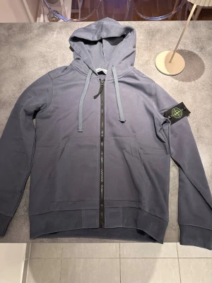 Blå hoodie från Stone Island - Snygg blå hoodie från Stone Island med dragkedja framtill och klassisk logotyp-patch på vänster ärm. Tröjan har huva med dragsko och ribbade muddar. Perfekt för en avslappnad och trendig stil.