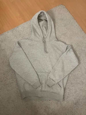 Grå hoodie från Bik Bok - Säljer en klassisk ljusgrå hoodie från Bik Bok med huva och dragsko. Hoodien har en stor magficka och ribbade muddar vid ärmslut och nederkant. Perfekt för en chill och avslappnad stil. Tillverkad i mjukt sweatshirtmaterial. Lite nopprig💕
