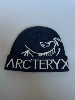 Mörkblå mössa från Arc'teryx - Säljer en mörkblå mössa från Arc'teryx med vit logga och text framtill. Mössan är stickad och har uppvikt kant. Insidan är ljus och mjuk för extra komfort. Perfekt för kalla dagar och snygg till streetwear-stilen.