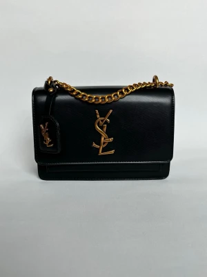 Svart axelväska från Saint Laurent - Snygg svart axelväska från Saint Laurent i slätt läder med guldfärgad kedja och ikoniska YSL-loggan framtill. Väskan har en stilren, rektangulär form och detaljer i guldmetall. Perfekt för dig som gillar lyxiga accessoarer med statement.