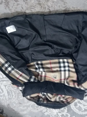 Burberry svart pufferjacka med huva - Svart pufferjacka från Burberry med ikoniskt beige, svart och rött rutigt foder. Jackan har huva, dragkedja och tryckknappar framtill samt två fickor med dragkedja. Materialet är vadderat och ger en varm känsla. Klassisk Burberry-detalj på bröstet och axeln.