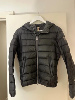 Svart jacka från Colmar - Snygg svart jacka från Colmar med huva och dragkedja framtill. Jackan har två sidofickor med dragkedja (ena är dock trasig, inget som syns) och Colmar-logga på ärmen. 