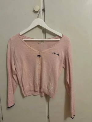 Ljusrosa Odd Molly kofta - Söt ljusrosa kofta från Odd Molly med v-ringning och knappar framtill. Liten broderad logga på bröstet och dekorativ lapp nedtill. Långärmad och i mjukt, tunt material. Perfekt att slänga över en topp för en chill look.