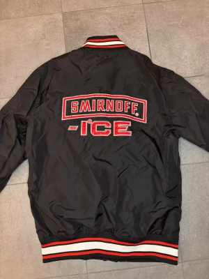 Svart bomberjacka Smirnoff Ice - Cool svart bomberjacka med stort Smirnoff Ice-tryck i rött och vitt på ryggen. Jackan har ribbade muddar och krage med röd, vit och svart rand. Materialet är glansigt och ger en sportig vibe, perfekt för dig som vill sticka ut.