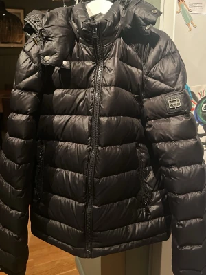 Svart pufferjacka från Bvaldi XXS - Snygg svart pufferjacka från Bvaldi i storlek XXS. Jackan har hög krage, avtagbar huva och två dragkedjefickor framtill. Puffig design med quiltade sektioner och en logga på ärmen. Perfekt för kalla dagar och riktigt varm. Använd 1 vinter. Nypris 3,500, priset är ej hugget i sten.