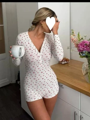Vit pyjamasbody med små rosa blommor - Supermysig vit pyjamasbody med lång ärm och korta ben, dekorerad med små rosa blommor. Djup v-ringning med knappar framtill. Perfekt för chillkvällar hemma.