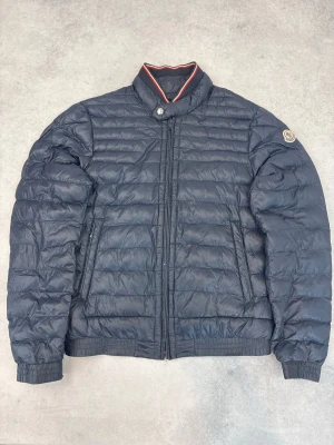 Mörkblå dunjacka från Moncler - Snygg mörkblå dunjacka från Moncler med quiltad design och ståkrage med rödvit detalj. Jackan har dragkedja framtill, två sidofickor och Moncler-logga på ärmen. Perfekt för kalla vinterdagar och riktigt stilren look.