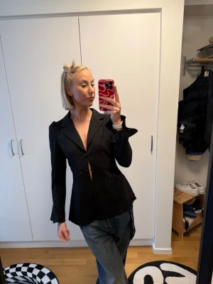 Svart kavaj från AÉLAN, storlek M - Snygg svart kavaj från AÉLAN med markerad midja och djup V-ringning. Kavajen har långa ärmar och en stilren, figurnära passform som ger en modern siluett. Perfekt för att lyfta vilken outfit som helst.