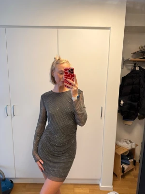 Glittrig silver kortklänning - Snygg kort klänning i silver med glittriga detaljer. Klänningen har lång ärm och figurnära passform, perfekt för dig som vill sticka ut. Materialet känns stretchigt och bekvämt. Ingen synlig logga eller märke.