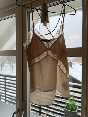 Beige spetslinnetopp med tunna band - Säljer en beige linnetopp med tunna axelband och vackra spetsdetaljer längs urringningen och framtill. Toppen är lätt transparent och har en feminin, luftig passform. Perfekt för dig som gillar en romantisk och trendig stil.