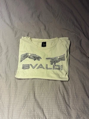 Bvaldi T-Shirt - Vit t-shirt från Bvaldi med glittriga strasstenar som formar två händer och texten 'BVALDI' på bröstet. Klassisk rund halsringning och korta ärmar. Perfekt för dig som gillar unika detaljer och lite extra bling.