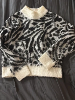 Zebramönstrad stickad tröja XS - Säljer en fluffig stickad tröja med zebramönster i svart och vitt. Tröjan har hög krage, breda ribbade muddar och nederkant. Perfekt för dig som vill ha en trendig och mysig look till hösten och vintern.