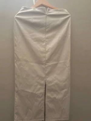 Beige pennkjol med slits bak - Stilren beige pennkjol i midi-längd med slits bak och dragkedja. Kjolen har en enkel, rak passform och är tillverkad i ett slätt tyg som ger en clean look. Perfekt att matcha med crop tops eller oversized tröjor för en trendig vibe.