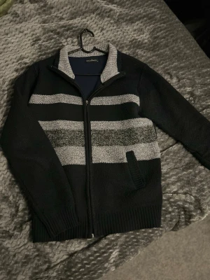 Svart ziptröja med gråa ränder Goldimen - Snygg svart ziptröja från Goldimen med breda gråa och ljusgråa ränder över bröstet. Tröjan har hög krage, ribbade muddar och dragkedja framtill. Perfekt för dig som gillar en stilren och enkel look.