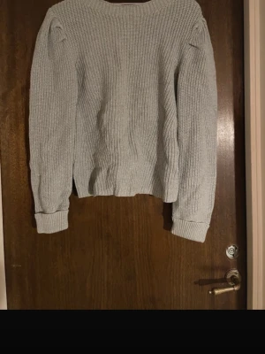 Ljusgrå stickad tröja med puffärm - Mysig ljusgrå stickad tröja med puffärmar och bred ribbad mudd vid ärmslut och nederkant. Tröjan har en klassisk rund halsringning och är tillverkad i ett grovstickat material som ger en avslappnad och trendig look.