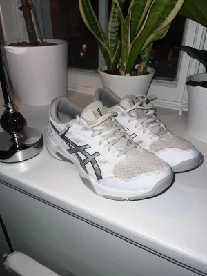 Vita Asics Gel-Rocket sneakers - Snygga vita Asics Gel-Rocket sneakers med grå och svarta detaljer. Skorna har en låg siluett, meshpaneler för bra ventilation och slitstarka syntetiska överdrag. Perfekta för dig som gillar sportig stil och komfort.