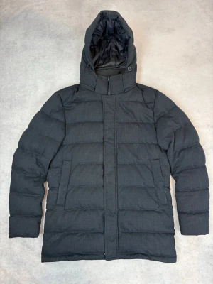 Svart pufferjacka från Herno - Stilren svart pufferjacka från Herno med quiltad design och avtagbar huva. Jackan har två sidofickor, hög krage och dragkedja med vindslå. Yttermaterialet är matt och känns slitstarkt, perfekt för kalla vinterdagar.