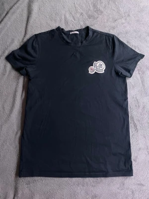 Svart Moncler t-shirt med logga - Snygg svart t-shirt från Moncler med två broderade loggor på bröstet. Klassisk rund halsringning och korta ärmar. Tillverkad i mjuk bomull som känns skön mot huden. Perfekt för dig som gillar stilrena plagg med exklusiv känsla.