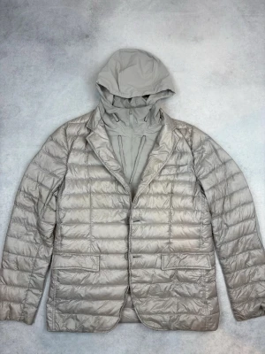 Beige dunjacka med huva från Herno - Snygg beige dunjacka från Herno med quiltad design och avtagbar huva. Jackan har lång ärm, dragkedja och knappar framtill samt två fickor med lock. Perfekt för kyliga dagar och ger en clean, modern look.