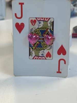 Rosa hjärtformade örhängen - Säljer ett par söta örhängen med hjärtformade hängen i genomskinlig rosa akryl. Örhängena har metallkrokar och hjärtana har en glansig finish med inslag av guldskimmer. Perfekta för dig som vill ha något gulligt och färgglatt i öronen.