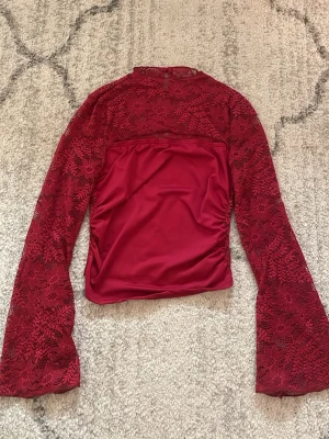 Röd spetsblus med utsvängda ärmar - Snygg röd topp i spets med långa utsvängda ärmar och rund hals. Toppen har ett genombrutet blommönster och en slät, fodrad kropp. Perfekt för dig som vill sticka ut med en trendig och elegant look.