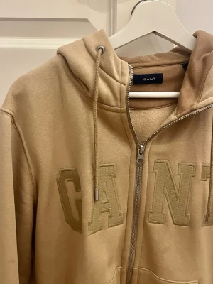 Beige hoodie med dragkedja från GANT - Snygg beige hoodie från GANT med dragkedja framtill och stora broderade bokstäver på bröstet. Hoodien har huva med dragsko och två fickor framtill. Tillverkad i mjukt bomullsmaterial och har ribbade muddar vid ärmslut och nederkant.