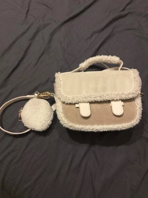 Söt liten väska🩷 - Söt handväska i beige och vitt med fluffiga detaljer och matchande axelrem. Väskan har ett litet fack framtill och ett gulligt, fluffigt myntfodral som hänger på remmen. Materialet är en mix av mockaimitation och teddy, perfekt för dig som gillar mysiga accessoarer.