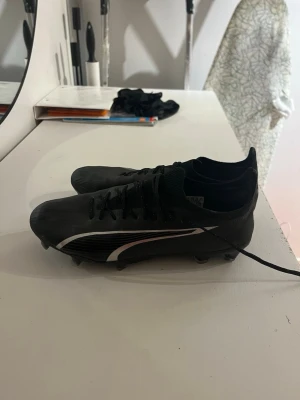 Svarta Puma Ultra fotbollsskor - Säljer ett par svarta Puma Ultra fotbollsskor med vit logga på sidan. Skorna har snörning och är tillverkade i ett lätt syntetmaterial för bra grepp och snabbhet på planen. Perfekta för dig som vill ha stilrena och snabba skor till match eller träning.