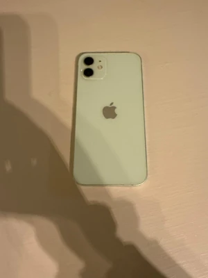Grön iPhone 12 med dubbla kameror - Snygg grön iPhone 12 med stilren design och rundade kanter. Baksidan är i glas och mobilen har två kameror på baksidan samt Apples logga i mitten. Skärmen är stor och täcker nästan hela framsidan med en liten notch upptill.