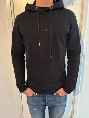 Les Deux hoodie   - Snygg svart hoodie från Les Deux med klassisk känguruficka och justerbar huva med snören. Diskret broderad logga på bröstet. Tillverkad i mjukt bomullsmaterial som känns skönt mot huden. Perfekt för en avslappnad och stilren look.