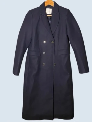 Ivy & Oak blå kappa, storlek 36 - Stilren mörkblå kappa med dubbelknäppning och klassisk krage. Lång modell med raka ärmar och två fickor framtill. Tillverkad i ullblandning för extra värme och snygg passform. Perfekt för kyliga dagar och ger en tidlös look.
