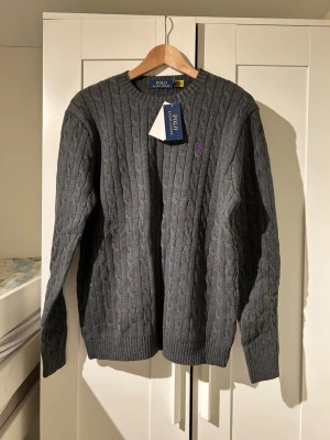 Grå kabelstickad tröja Polo Ralph Lauren - Kabelstickad grå tröja från Polo Ralph Lauren med rund halsringning och ribbade muddar. Tröjan har en lila broderad logga på bröstet och är tillverkad i mjukt material. Perfekt för dig som gillar klassisk och stilren design. Helt ny oanvänd. Priset kan diskuteras Storlek M 