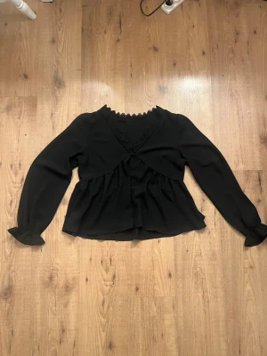 Svart volangblus med spetsdetalj - Svart blus i luftig chiffong med volang nedtill och spetskantad V-ringning. Långa ärmar med lätt puff och volang vid ärmslut. Perfekt för en trendig och feminin look.