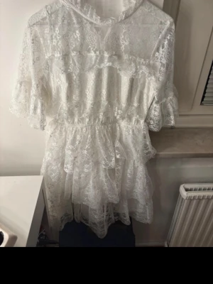 Studentklänning  - Studentklänning i spets. Romantisk vit spetsklänning från Bubbleroom med volangdetaljer och markerad midja. Klänningen har kort längd, genombruten spets och korta ärmar med volang. Perfekt för sommarens alla tillfällen och ger en söt och feminin look.