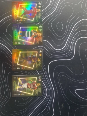 Fyra glittriga fotbollssamlarbilder - Säljer fyra glittriga samlarkort med fotbollsspelare från Champions League. Korten har färgstarka holografiska effekter och visar spelare i matchställ från olika lag. Perfekt för dig som gillar att samla på fotbollskort eller vill utöka din samling.