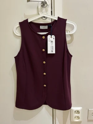  Ny Vinröd ärmlös topp Holly & Whyte - Snygg vinröd ärmlös topp från Holly & Whyte med rund halsringning och dekorativa guldknappar framtill. Toppen har en stilren och figurnära passform, perfekt att matcha med både jeans och kjol. Materialet känns mjukt och bekvämt.