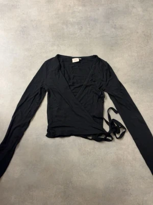 Svart omlottopp med långa ärmar - Snygg svart omlottopp med långa ärmar och knytband i midjan. Toppen har en djup v-ringning och croppad passform som ger en trendig look. Perfekt att styla med höga jeans eller kjol.