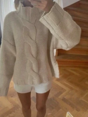 Beige kabelstickad polotröja med prislapp  - Säljer en beige, chunky kabelstickad polotröja med breda ärmar och hög krage. Tröjan har en oversized passform och grovstickat mönster framtill. Perfekt för dig som gillar mysiga och trendiga plagg till hösten och vintern.