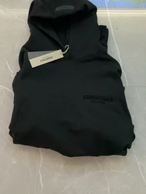 Svart hoodie från Essentials - Säljer en svart hoodie från Essentials Fear of God med diskret logga på bröstet. Hoodien har en klassisk huva och dragsko, samt en stilren look utan synliga fickor framtill. Perfekt för dig som gillar minimalistisk streetwear.
