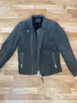 Grå mocka från Jofama - Snygg grå skinnjacka från Jofama med klassisk bikerstil. Jackan har sned dragkedja framtill, två dragkedjeförsedda fickor och coola detaljer. Perfekt för dig som vill ha en stilren och tidlös look.