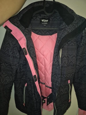 Barn svart rosa vinter jacka  med mönster från Woxer från kappahl stl 164 - Mönstrad svart ,rosa  vinterjacka från Woxer med rosa quiltat foder och detaljer. Jackan har dragkedja, huva och justerbara snören i midjan. Snygg grafisk design och praktiska fickor med dragkedja. Perfekt för dig som vill ha en unik och funktionell jacka. Stl 164