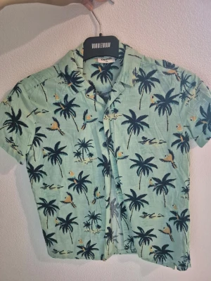  Barn Grön skjorta med palm- och papegojmönster stl 134 - Kortärmad skjorta från Kaporal i ljusgrönt med tropiskt mönster av palmer och papegojor. Skjortan har klassisk krage och knappar framtill. Perfekt för dig som gillar färgglada och somriga plagg.