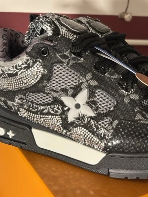 Louis Vuitton svarta sneakers med kristaller - Unika svarta Louis Vuitton sneakers med glittrande kristalldetaljer och ikoniskt monogrammönster. Skon har en låg siluett, svarta snören och kontrasterande vita detaljer på sulan. Materialet är en mix av textil och syntet med exklusiva LV-detaljer.