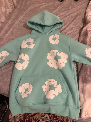 Denim tears hoodie med vita molntryck - Cool denim tears hoodie med stora vita molnliknande tryck över hela tröjan. Hoodien har en klassisk huva och en stor magficka framtill. Perfekt för dig som gillar unika och färgglada plagg. Materialet känns mjukt och skönt mot huden. Helt ny aldrig använd