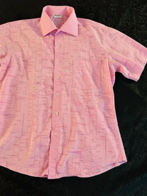 Rosa kortärmad skjorta från Mixers  - Säljer en rosa skjorta från Mixers  med abstrakt mönster i lila toner. Skjortan har korta ärmar, klassisk krage och knäppning framtill. Perfekt för dig som vill sticka ut lite extra med en unik look.