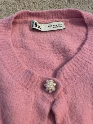 Rosa kofta med blomknappar från ZARA - Supersöt rosa kofta från ZARA med långa ärmar och rund hals. Koftan har unika blommiga knappar som ger en gullig touch. Mjuk och skön, perfekt att slänga på sig när det är lite kyligt ute.