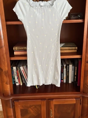 Ljusblå kortklänning med blommor - Söt ljusblå kortklänning med små vita och gula blommor, korta ärmar och volangkant upptill och nedtill. Perfekt för sommarens varma dagar och har en slim passform.